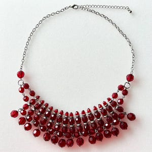 Op de afbeelding: Een rode kralenketting met een zilverkleurige ketting. De ketting heeft meerdere strengen van facetgeslepen rode kralen, waardoor een watervaleffect ontstaat. De kralen variëren in grootte, wat visuele interesse aan de sieraden toevoegt.