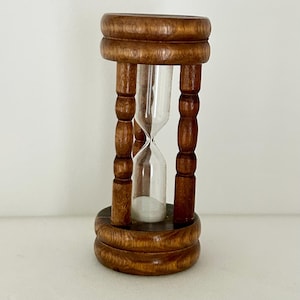 May include: A wooden hourglass featuring a clear glass centre filled with white sand. The frame consists of a circular top and base connected by four turned, spindle-style pillars with a polished brown finish and visible wood grain texture.