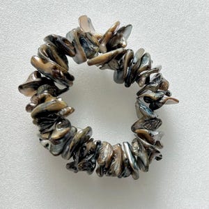 Puede incluir: Un brazalete hecho de piezas de concha iridiscente, marrón y negra. El brazalete está hecho de muchas piezas pequeñas e irregulares de concha que se ensartan para formar un círculo.