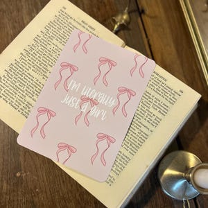 Puede incluir: Una tarjeta rosa con un patrón repetido de lazos rosas. La tarjeta tiene el texto "I'm literally just a girl" en letras negras.
