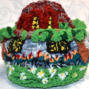 May include: A handmade, colorful teapot cozy shaped like a whimsical house. The cozy features a red roof, green foliage, and windows with yellow accents. The base is green with white and yellow flower details.
