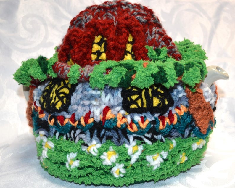 May include: A handmade, colorful teapot cozy shaped like a whimsical house. The cozy features a red roof, green foliage, and windows with yellow accents. The base is green with white and yellow flower details.