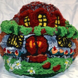 May include: A whimsical, hand-knitted tea cozy shaped like a colorful cottage. The cozy features a red roof, yellow-lit windows, a green lawn with white flowers, and a brown door. The cozy is designed to fit a standard teapot.
