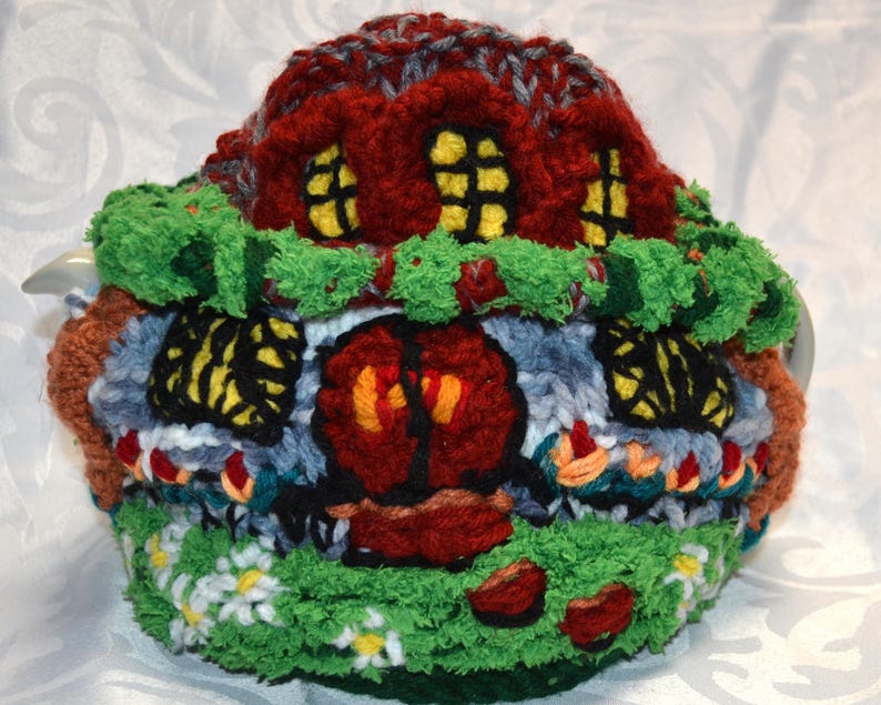 May include: A whimsical, hand-knitted tea cozy shaped like a colorful cottage. The cozy features a red roof, yellow-lit windows, a green lawn with white flowers, and a brown door. The cozy is designed to fit a standard teapot.