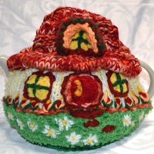 May include: A handmade, knitted teapot cozy shaped like a whimsical house. The cozy features a red roof, yellow windows, and a green base with white daisy accents. The cozy is designed to fit a standard teapot.