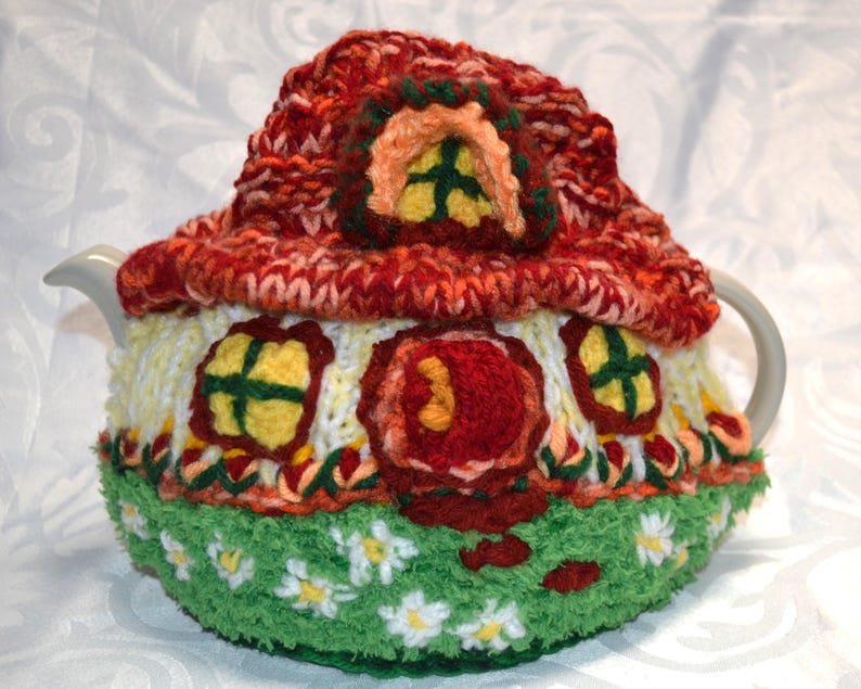 May include: A handmade, knitted teapot cozy shaped like a whimsical house. The cozy features a red roof, yellow windows, and a green base with white daisy accents. The cozy is designed to fit a standard teapot.