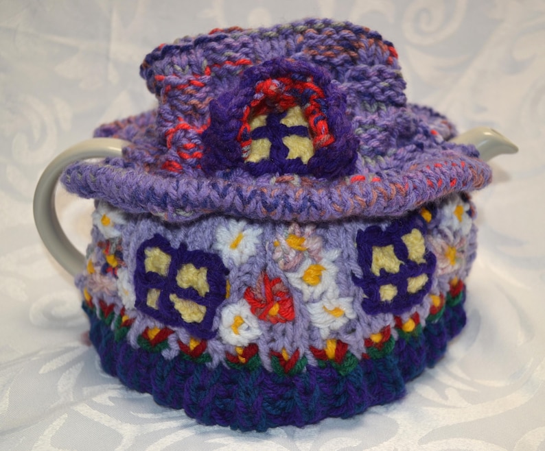 May include: A whimsical, hand-crocheted tea cozy in shades of purple, blue, red, and yellow. The cozy is designed to resemble a fairy house, complete with windows and a roof. The cozy is designed to fit a standard size teapot.