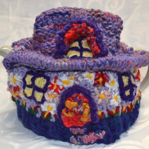 May include: A whimsical, handmade crochet tea cozy in shades of purple, red, yellow, and white. The cozy is designed to resemble a fairy house, complete with a roof, windows, and a door. The cozy is designed to fit a teapot.