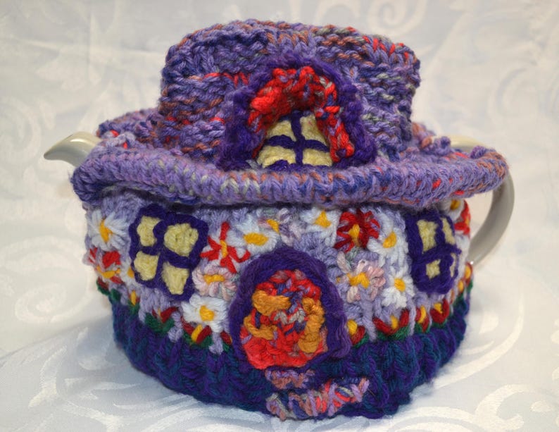 May include: A whimsical, handmade crochet tea cozy in shades of purple, red, yellow, and white. The cozy is designed to resemble a fairy house, complete with a roof, windows, and a door. The cozy is designed to fit a teapot.