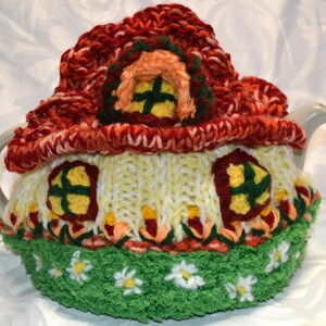 May include: A whimsical, hand-knitted tea cozy shaped like a cottage. The cozy features a red roof, yellow walls with windows, and a green base with white daisy accents. The cozy is designed to fit a standard teapot.