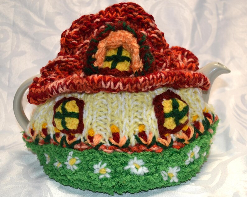 May include: A whimsical, hand-knitted tea cozy shaped like a cottage. The cozy features a red roof, yellow walls with windows, and a green base with white daisy accents. The cozy is designed to fit a standard teapot.
