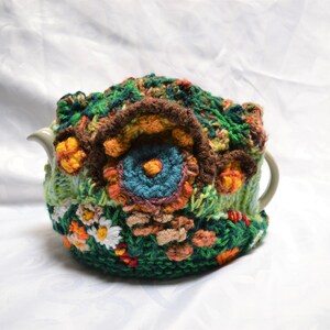May include: A handmade, crocheted tea cozy in shades of green, brown, orange, and blue. The cozy features a floral design with detailed stitching and a textured appearance, designed to fit a teapot.