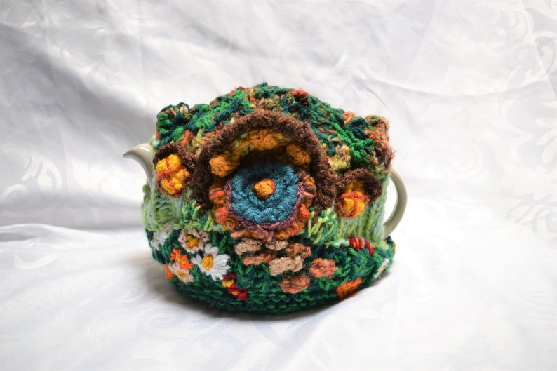 May include: A handmade, crocheted tea cozy in shades of green, brown, orange, and blue. The cozy features a floral design with detailed stitching and a textured appearance, designed to fit a teapot.