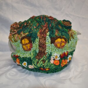 May include: A whimsical, hand-crocheted tea cozy in the shape of a fairy house. The cozy features a tree, two windows with yellow light, and colorful flowers. The color palette includes green, brown, yellow, orange, and white.