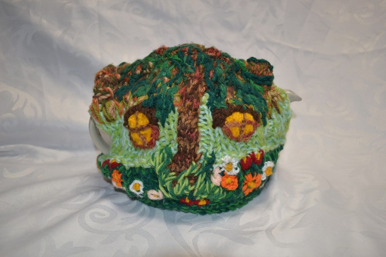 May include: A whimsical, hand-crocheted tea cozy in the shape of a fairy house. The cozy features a tree, two windows with yellow light, and colorful flowers. The color palette includes green, brown, yellow, orange, and white.