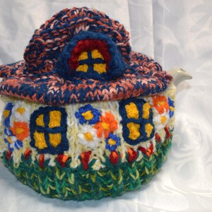 May include: A whimsical, hand-knitted teapot cozy shaped like a house. The cozy features a blue and orange roof, yellow window details, and a green base. The cozy is designed to fit a standard teapot.