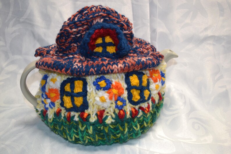 May include: A whimsical, hand-knitted teapot cozy shaped like a house. The cozy features a blue and orange roof, yellow window details, and a green base. The cozy is designed to fit a standard teapot.