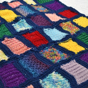 May include: A colourful crocheted blanket with squares of different colours and textures. The blanket is made with a variety of yarns, including some with a fluffy texture. The squares are sewn together with a dark blue yarn, creating a patchwork effect.