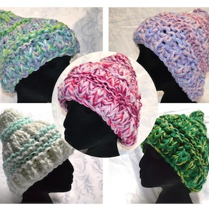 May include: A collection of five chunky knit hats in a variety of colours, displayed on black mannequin heads. The hats are hand-knitted with a thick yarn, creating a textured appearance. Colours include green, blue, purple, pink, and white.