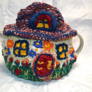 May include: A whimsical, hand-knitted teapot cozy shaped like a colorful cottage. The cozy features a blue roof, yellow windows, and a red door, with floral and grass details. The cozy is designed to fit a standard teapot.