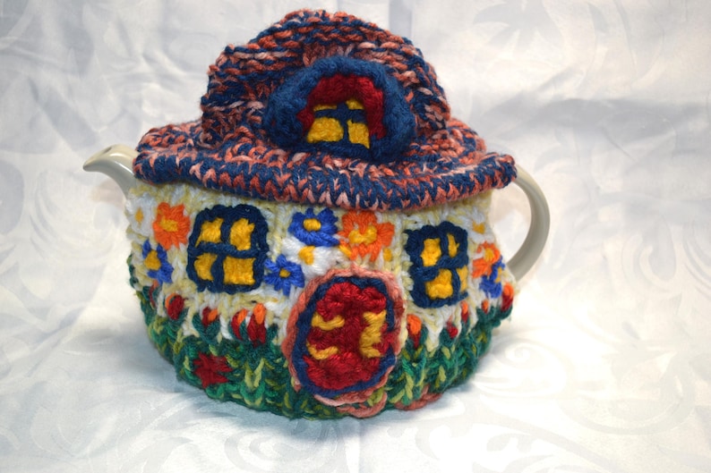 May include: A whimsical, hand-knitted teapot cozy shaped like a colorful cottage. The cozy features a blue roof, yellow windows, and a red door, with floral and grass details. The cozy is designed to fit a standard teapot.
