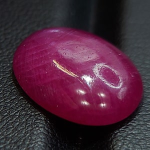 May include: A vibrant, oval-shaped ruby gemstone with a deep red hue. The stone is polished and has a smooth, glossy finish.