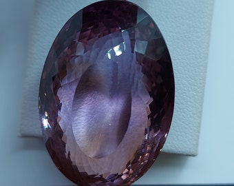May include: A large, oval-shaped, faceted amethyst gemstone with a purple and pink hue. The gemstone is set against a white background.