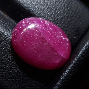 Peut inclure: Une pierre précieuse ovale de couleur rose foncé avec une surface lisse et polie. La pierre précieuse a un léger éclat et est placée sur un fond noir.