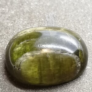 May include: A smooth, oval-shaped green gemstone with a dark green, almost black, center. The stone has a glossy finish and appears to be translucent.