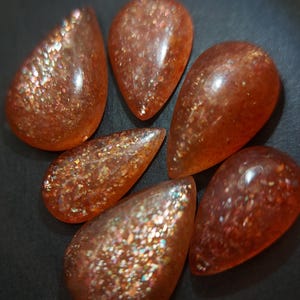May include: Six teardrop-shaped sunstone cabochons with a shimmering, iridescent finish. The stones are a mix of orange, brown, and gold colors.