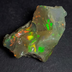 Op de afbeelding: Een ruwe, ongeslepen opaal edelsteen met levendige kleuren. De steen vertoont flitsen van groen, oranje en geel in een doorschijnende, melkachtige basis. De opaal is onregelmatig van vorm en toont zijn natuurlijke, ruwe vorm tegen een donkere achtergrond.