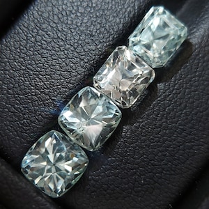 May include: Four clear, faceted, square-shaped gemstones arranged in a row on a black leather surface. The gemstones are different sizes and have a light blue hue.