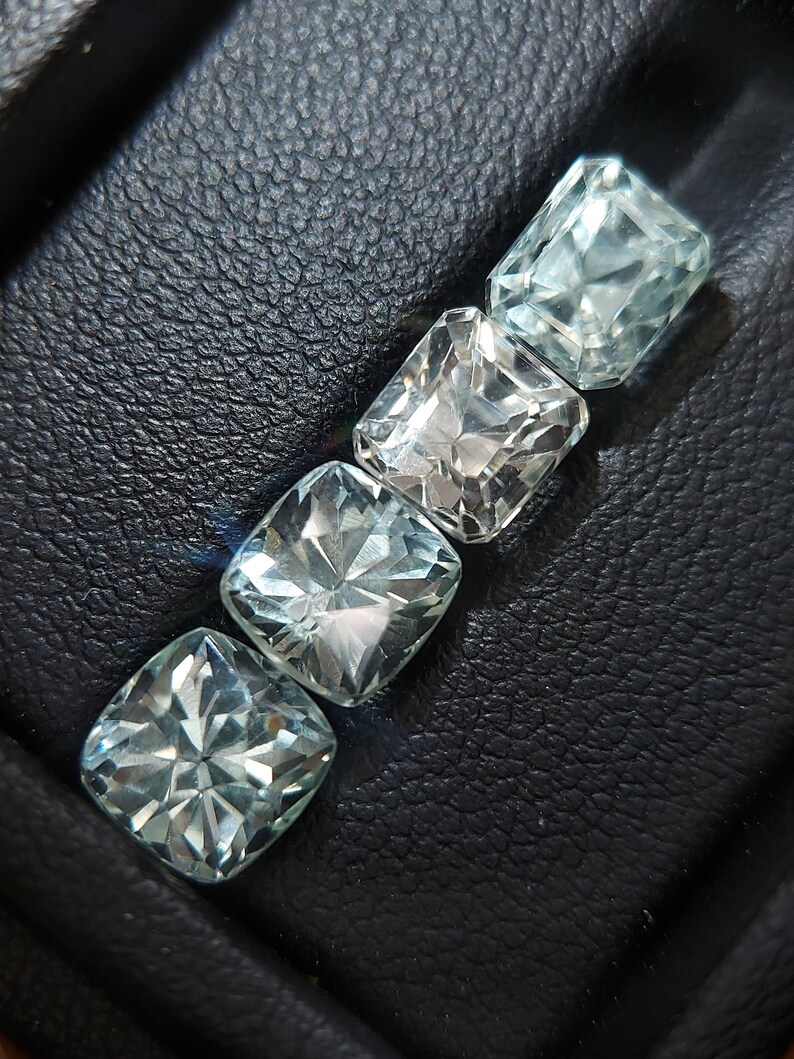 May include: Four clear, faceted, square-shaped gemstones arranged in a row on a black leather surface. The gemstones are different sizes and have a light blue hue.