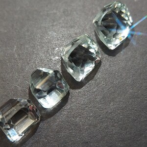 May include: Four clear, faceted, light green gemstones, cut in a square shape, arranged in a row on a dark gray surface.