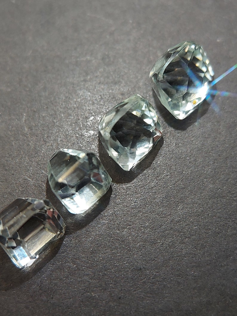 May include: Four clear, faceted, light green gemstones, cut in a square shape, arranged in a row on a dark gray surface.