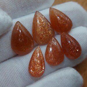 May include: Six teardrop-shaped sunstone cabochons in shades of orange and brown with gold flecks. The stones are arranged in a semi-circle on a white surface.