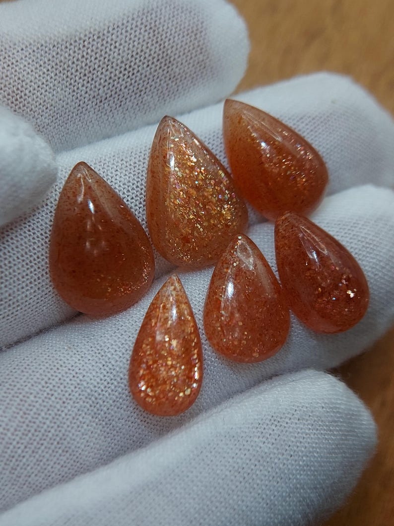 May include: Six teardrop-shaped sunstone cabochons in shades of orange and brown with gold flecks. The stones are arranged in a semi-circle on a white surface.