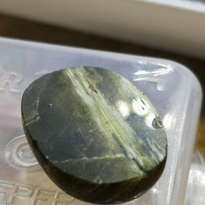 May include: A smooth, oval-shaped green and white gemstone with a striated pattern. The stone is polished and has a glossy finish.