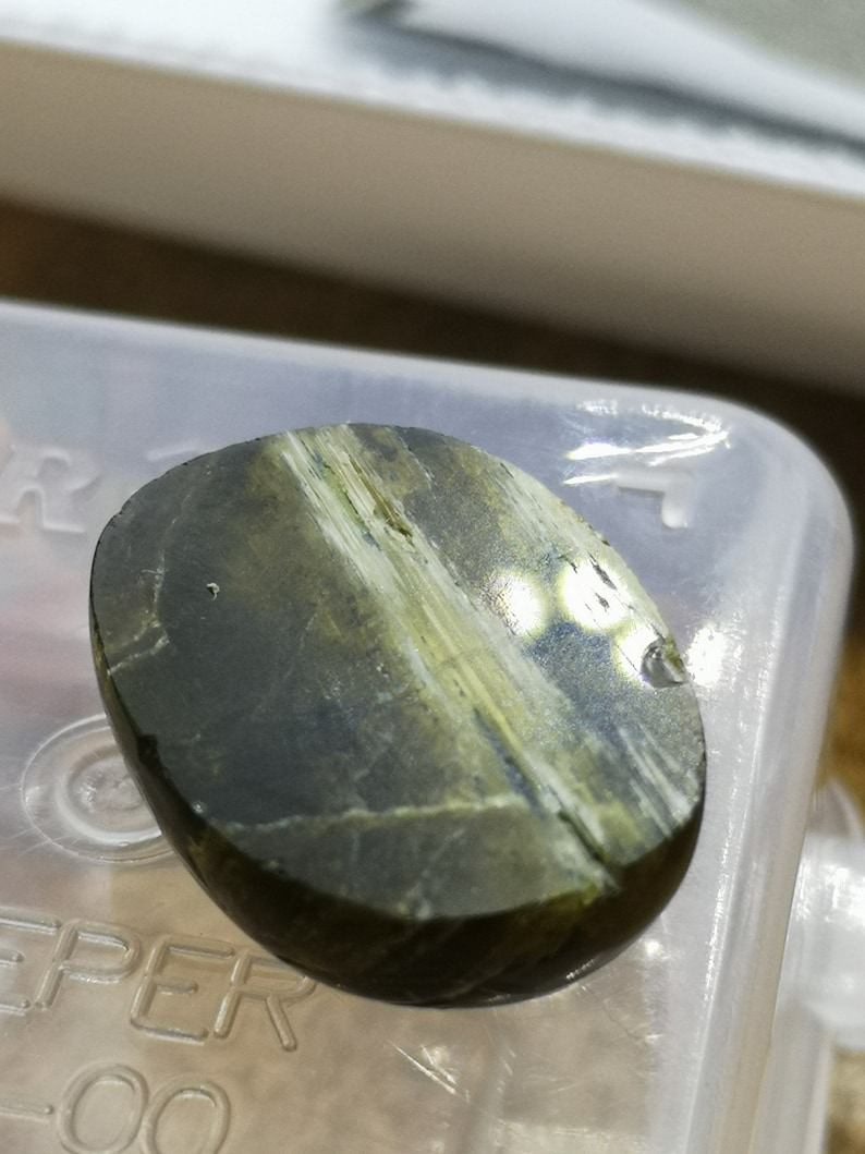 May include: A smooth, oval-shaped green and white gemstone with a striated pattern. The stone is polished and has a glossy finish.