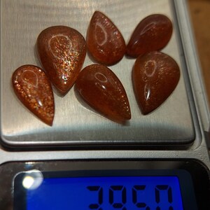 May include: Six teardrop-shaped, orange-brown sunstone gemstones on a digital scale. The scale reads 39.50 ct.