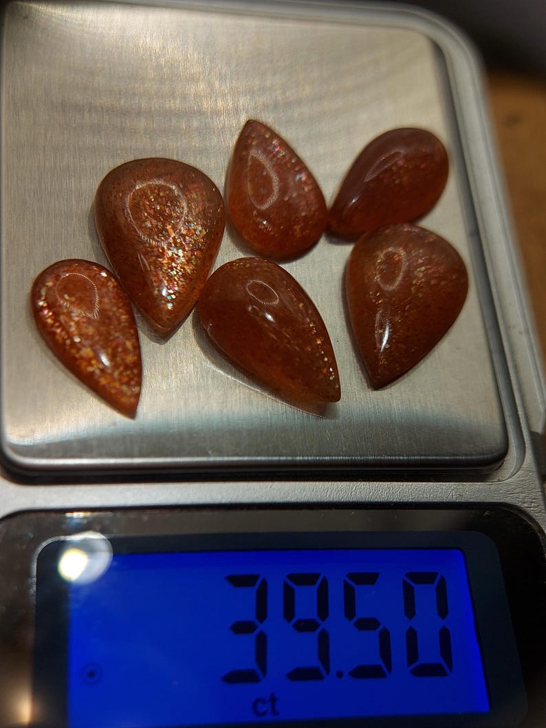 May include: Six teardrop-shaped, orange-brown sunstone gemstones on a digital scale. The scale reads 39.50 ct.