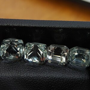 May include: Four green gemstones in a square shape are displayed in a black leather box. The gemstones are cut and polished, and they have a clear, glassy appearance.