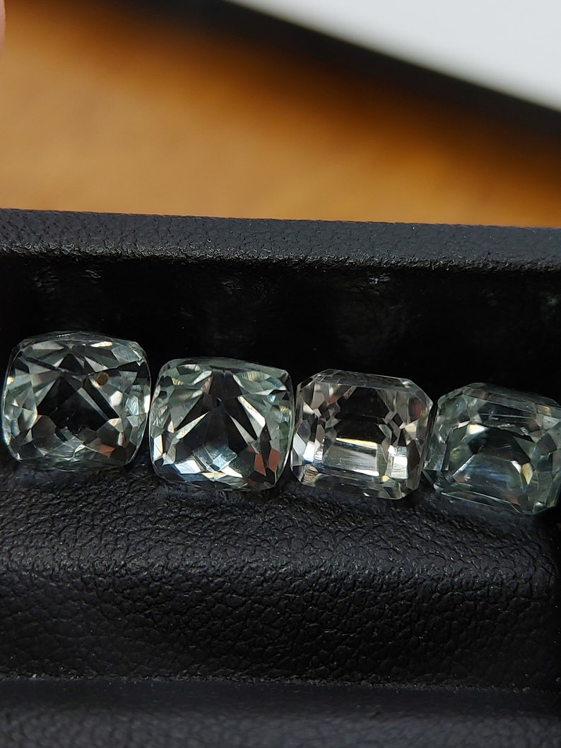 May include: Four green gemstones in a square shape are displayed in a black leather box. The gemstones are cut and polished, and they have a clear, glassy appearance.