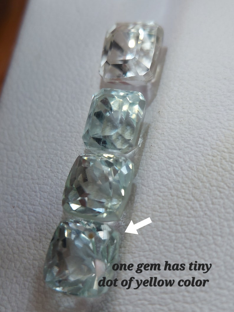 May include: Four light blue aquamarine gemstones, one with a tiny yellow dot, arranged vertically on a white surface.