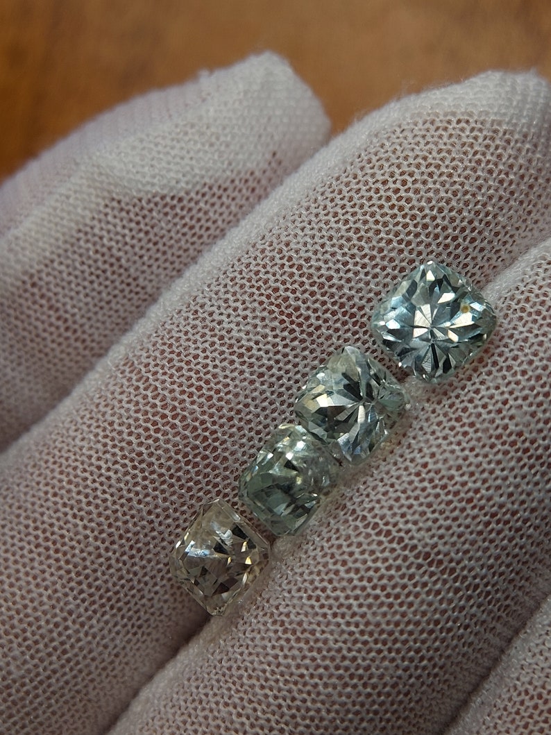 May include: Four faceted, clear, light green gemstones with a square shape. The gemstones are arranged in a row, with the largest gemstone at the top and the smallest gemstone at the bottom.