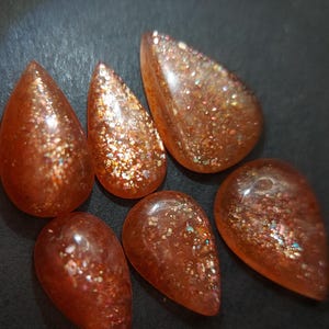May include: Six teardrop-shaped sunstone cabochons with a shimmering, iridescent finish. The stones are a mix of orange, red, and gold.