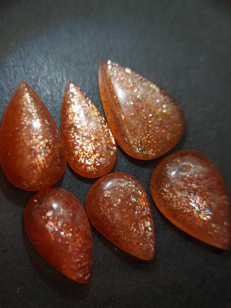 May include: Six teardrop-shaped sunstone cabochons with a shimmering, iridescent finish. The stones are a mix of orange, red, and gold.