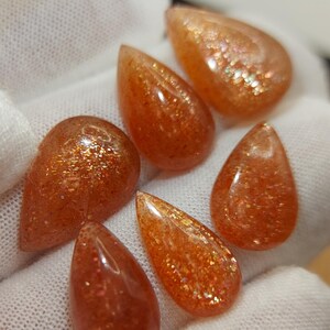 May include: Six teardrop-shaped sunstone cabochons with a shimmering, iridescent finish. The stones are a mix of orange, red, and gold hues.