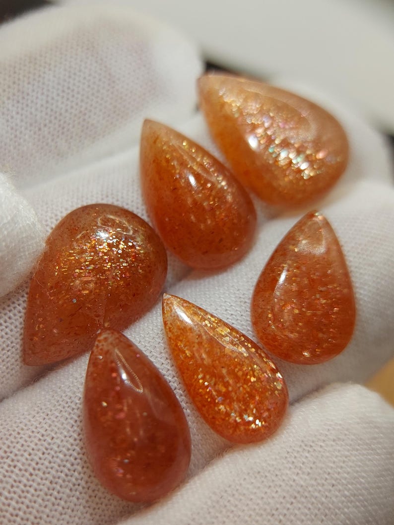 May include: Six teardrop-shaped sunstone cabochons with a shimmering, iridescent finish. The stones are a mix of orange, red, and gold hues.