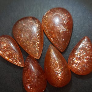 May include: Six teardrop-shaped sunstone cabochons with a shimmering, iridescent finish. The stones are a mix of orange and red hues.